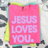 Jesus Loves You LAVENDER DTF Heat Transfer, pink T-shirt with purple text, stacked with green and turquoise shirts on a patterned surface.