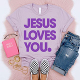 Jesus Loves You PURPLE DTF Heat Transfer, purple T-shirt with bold lettering, casual jeans, checkered slip-on shoes, Holy Bible, and jewelry arranged on a pink fluffy surface.