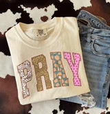 PRAY Patchwork DTF Transfer | Fall Faith Design with Floral & Pumpkin Print Letters, a beige sweatshirt with colorful patchwork-style 'PRAY' lettering and a pair of distressed blue jeans, on a patterned textile background.