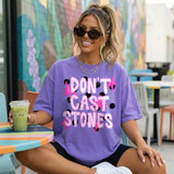 Don't Cast Stones DTF Transfer, retro bold lettering faith design on a woman's purple T-shirt, modern fashion style, outdoor cafe setting.