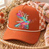 Dove Day DTF transfer featuring a whimsical patchwork-style dove in flight made of mixed bright patterns with bubble-letter DOVE and DAY text Shown on a Burnt orange front panel with cream rope and orange/ivory checkered mesh back trucker hat.