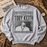 Toby Keith DTF transfer designed as a vintage newspaper front page with distressed photo and 'Should've Been a Cowboy - As Good As I Once Was' headline Shown on a Sport Grey crewneck sweatshirt.