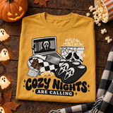 Cozy Nights Are Calling Halloween DTF transfer with laptop playing horror movie, checkered blanket, chocolate bar, skull popcorn bucket, and ghost-face slippers Shown on a Mustard t-shirt.