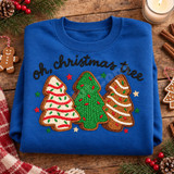Oh Christmas Tree DTF transfer with three crocheted-look Christmas tree cookie designs in red and white, green, and gingerbread icing styles, surrounded by stars, under black brush-script lettering Shown on a Sapphire crewneck sweatshirt.
