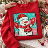 Cheerful waving snowman DTF transfer wearing a red Santa hat, mittens, and scarf on a teal and pink splatter background with snowflakes and glitter dots Shown on a Red crewneck sweatshirt.