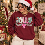 JOLLY DTF transfer in polka-dot bubble letters with pine garland, glitter berries, and a pink bow Shown on a Garnet crewneck sweatshirt.