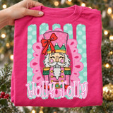Holly Jolly nutcracker DTF transfer with pink and green nutcracker face, red bow, gold crown, in a pink scalloped frame over a mint polka-dot striped background with bold bubble lettering Shown on a Crunchberry t-shirt.