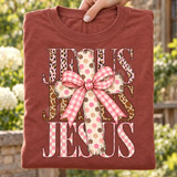 JESUS DTF transfer with pink leopard, giraffe print, and pink checkerboard lettering, layered with a wood-grain polka dot cross and pink gingham bow Shown on a Brick t-shirt.
