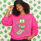 Goofy Green Monster Halloween DTF Transfer | Funny Retro Monster Face Design, smiling model wearing a bright pink sweatshirt with a large vintage-style green monster face graphic, against a Halloween background of green ghosts and jack-o’-lanterns.