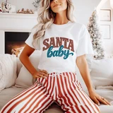 Santa Baby DTF Transfer | Retro Christmas Design, retro red and teal “Santa Baby” graphic displayed on a white T-shirt worn with red-and-white striped pants in a cozy Christmas living room