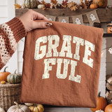 Grateful DTF Transfer | Distressed Collegiate Fall Design, distressed cream collegiate “Grateful” lettering displayed on a rust-colored shirt in a cozy autumn setting with pumpkins and fall decorations