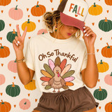 Oh So Thankful Turkey DTF Transfer | Fall Thanksgiving Design, colorful turkey with patterned feathers and “Oh So Thankful” lettering on a cream T-shirt, modeled against a fall pumpkin background
