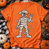 Rock On Mummy Halloween DTF Transfer | Funny Cartoon Mummy Design, playful bandaged cartoon mummy making a rock hand sign printed on an orange T-shirt with colorful splatters, surrounded by Halloween pumpkins and decor.
