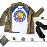 Made in the 80s Rainbow Brite Sublimation Transfer, graphic design featuring a colorful cartoon girl with stars, on a T-shirt with a navy blue and white sleeves, laid on a white wooden surface with a khaki jacket, black boots, small potted plants, and a magazine.