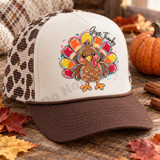 Give Thanks Turkey DTF Transfer | Cute Thanksgiving Fall Design, colorful cartoon turkey with “Give Thanks” lettering displayed on a cream and brown trucker hat in a festive autumn setting