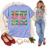 Three pink, teal and gold nutcracker soldiers DTF transfer holding a candy cane, drum and Christmas tree, framed by pine garland and holly Shown on a Hydrangea t-shirt.
