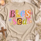 Blessed DTF transfer with scrapbook-style polka-dot, plaid, and star paper cutout letters and a pink ribbon bow Shown on a Sandstone t-shirt.