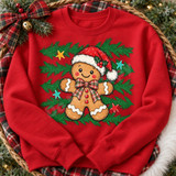 Gingerbread man DTF transfer wearing a sparkly Santa hat and plaid bow, surrounded by pine greenery and colorful stars Shown on a Cardinal Red crewneck sweatshirt.