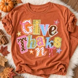 Give Thanks scrapbook-style word art DTF transfer with mixed patterned paper letters, pink ribbon bows, and sparkly stars Shown on a Terracotta t-shirt.