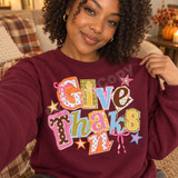 Give Thanks scrapbook-style word art DTF transfer with mixed patterned paper letters, pink ribbon bows, and sparkly stars Shown on a Maroon crewneck sweatshirt.