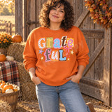 Grateful scrapbook-style word art DTF transfer with mixed-pattern paper-cut letters, pink ribbon bow, and stars in autumn pastel colors Shown on a Orange crewneck sweatshirt.