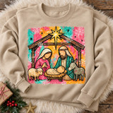 Folk-art nativity scene DTF transfer with Mary, Joseph, baby Jesus, sheep and donkey in a wooden stable under the Star of Bethlehem, on a splattered pink, orange and teal background with snowflakes Shown on a Sand crewneck sweatshirt.