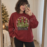 Peace on Earth Christmas DTF transfer with three folk-art green trees, gold stars, and red swirl ornaments inside a hand-brushed black circle frame Shown on a Antique Cherry Red crewneck sweatshirt.