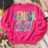 Senior 2027 DTF transfer in hand-painted graffiti-style rainbow lettering with black outlines and scattered doodle hearts Shown on a Heliconia crewneck sweatshirt.