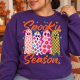 Spooky Season DTF transfer featuring four patterned ghosts in pink hearts, rainbow polka dots, floral, and checkerboard designs with orange script lettering and Halloween doodles Shown on a Purple crewneck sweatshirt.