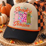 Spooky Season DTF transfer featuring four patterned ghosts in pink hearts, rainbow polka dots, floral, and checkerboard designs with orange script lettering and Halloween doodles Shown on a Khaki front panel with black bill and orange rope, black and orange checkered mesh back trucker hat.
