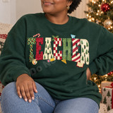 Frosted cookie-style TEACHER lettering DTF transfer with leopard print, Christmas trees, candy stripes and checkerboard patterns, surrounded by a candy cane, holly, paint palette, pencil, ruler and book icons Shown on a Forest crewneck sweatshirt.
