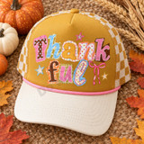 Thankful DTF transfer in whimsical torn-paper collage lettering with mismatched patterns, pink ribbon bow, and hand-drawn stars Shown on a mustard gold front panel with cream bill and cream-and-mustard checkered mesh back, pink rope trucker hat.