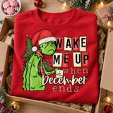 Grumpy green Grinch-inspired character in a Santa hat and candy-striped tie holding an 'I Hate Christmas' mug beside a decorated tree, with bold text reading 'Wake Me Up When December Ends' DTF transfer design Shown on a Red crewneck sweatshirt.