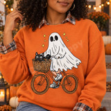 Cartoon ghost riding an orange bicycle with a black cat in the front basket and yellow sparkle stars, DTF transfer on a yellow t-shirt Shown on a Orange crewneck sweatshirt.