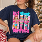 Nut About Christmas DTF transfer featuring three whimsical pink and turquoise nutcracker soldiers with crowns, a drum, candy cane, and tiny Christmas tree on pink polka-dot stripes Shown on a True Navy t-shirt.