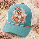 Whimsical pink and teal nutcracker head DTF transfer with gold jeweled details, pearl trim, florals, hearts, and watercolor splatters Shown on a dusty teal front panel, blush pink rope, white mesh back trucker hat.