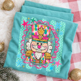 Whimsical nutcracker DTF transfer in pink, teal, and gold painterly style, framed by a pink scalloped oval with stars and snowflakes on a mint striped background Shown on a Island Reef t-shirt.