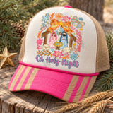 Oh Holy Night nativity DTF transfer with Mary, Joseph, baby Jesus, angel and sheep framed by an orange floral bow, gold star and pastel flowers, pink script lettering below Shown on a Ivory front panel with pink and gold striped bill and tan mesh back trucker hat.