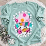 Colorful snowman face DTF transfer with polka-dot striped hat, carrot nose, pink cheeks, and winking eyes framed by a fringed border Shown on a Seafoam t-shirt.