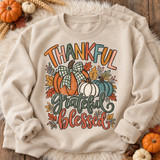 Thankful Grateful Blessed DTF transfer with hand-drawn orange, cream, and teal pumpkin trio, plaid bow, autumn leaves, and berries Shown on a Sand crewneck sweatshirt.