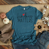 Kindness Matters Adult Screen Print Heat Transfer, a teal T-shirt with the phrase 'kindness MATTERS' and a red heart graphic, laid out on a rustic wooden surface with fall decor including pumpkins, cotton stems, a sunflower, and jeans.