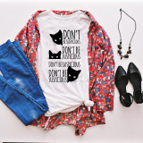 Don't be Suspicious Cat Sublimation Transfer, white t-shirt with black cat faces and the phrase 'Don't Be Suspicious' printed on it, styled with blue jeans, a colorful floral shirt, black shoes, necklace, and accessories.