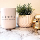 Find Your Motivation Sublimation Transfer, motivational phrase on a white mug with black floral illustrations, placed next to a green plant in a white pot and a shiny gold skull decor on marble surface.