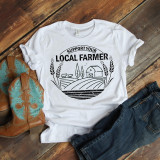 Support your local farmer sublimation transfer, printed on a white T-shirt with a farm scene design, next to cowboy boots and denim jeans on a wooden surface.