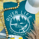 River Life Cuz Beaches Be Salty Screen Print Transfer, close-up of a turquoise t-shirt with a white graphic of trees, beach, and kayak, accompanied by a white cap, wooden bead necklace, and dried grass.