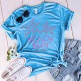 Beach Bum Screen Print Heat Transfer, light blue t-shirt with pink palm trees and text design, casual outfit with denim shorts and white sneakers, sunglasses and flowers accessories.