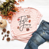 Fall for Jesus He Never Leaves Screen Print Transfer, pink T-shirt with autumn leaves and inspirational message on white wooden surface, with jeans, pumpkins, berries, and pinecones nearby.