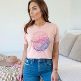 Adventure Awaits Watercolor Screen Print Heat Transfer, a woman wearing a pink T-shirt featuring a watercolor landscape with mountains, trees, and a lake, with the text "Adventure Awaits" design.