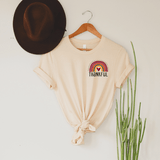 Thankful Rainbow HAT/POCKET Screen Print Transfer on a beige T-shirt with a knot at the bottom, hanging on a wooden hanger, with a brown hat and green plant nearby.