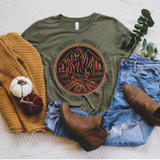Sweaters Hayrides Boots Pumpkins Screen Print Transfer, flat lay of casual fall-themed outfit with graphic T-shirt featuring a campfire design surrounded by text, paired with jeans, brown ankle boots, a mustard sweater, and small pumpkins, with greenery in the background.